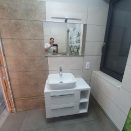 OTGlazurnik Małopolska Oskar Tracz - Nowoczesna łazienka w trakcie remontu, widoczna biała umywalka z szafką, beżowe płytki na ścianach, czarne okno, fragment ściany w surowym stanie, osoba robiąca zdjęcie w lustrze.