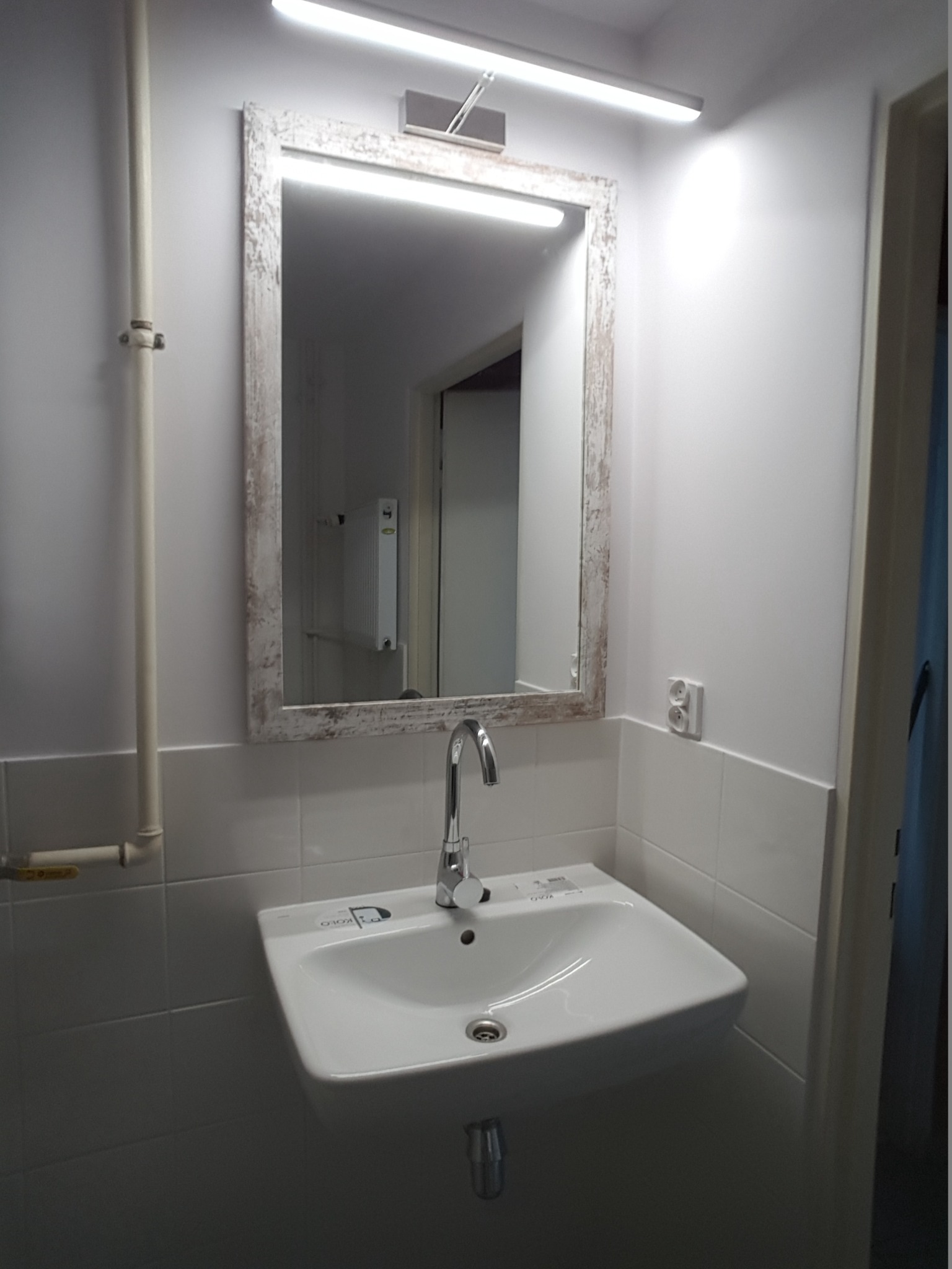 Biała umywalka z chromowaną baterią zamontowana na tle ściany z białych płytek, nad nią lustro w ozdobnej, postarzanej ramie i oświetlenie LED, widoczne elementy instalacji wodnej i elektrycznej.