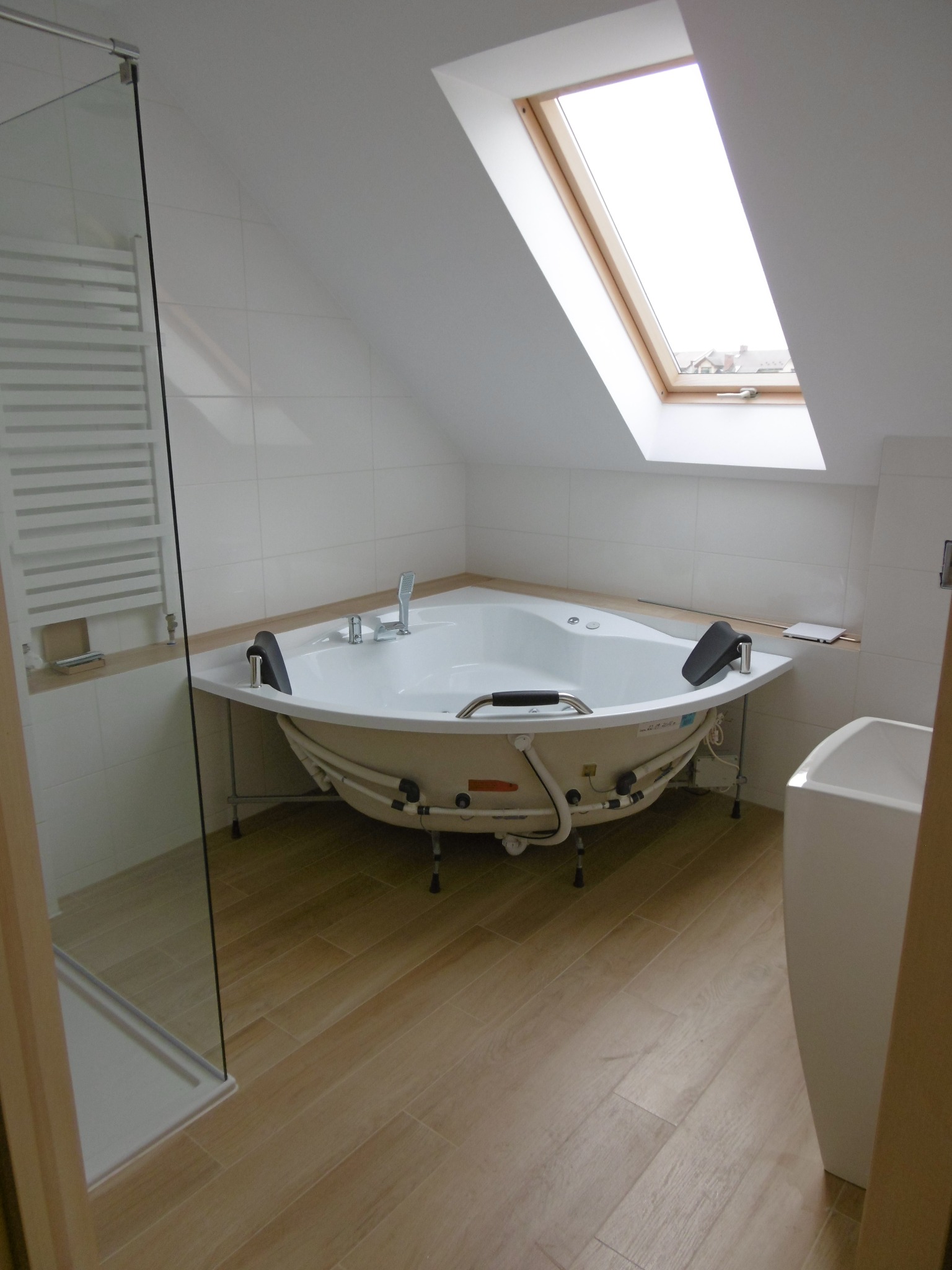 Nowoczesna łazienka na poddaszu z narożną wanną z hydromasażem, oknem dachowym i jasnymi płytkami imitującymi drewno na podłodze.