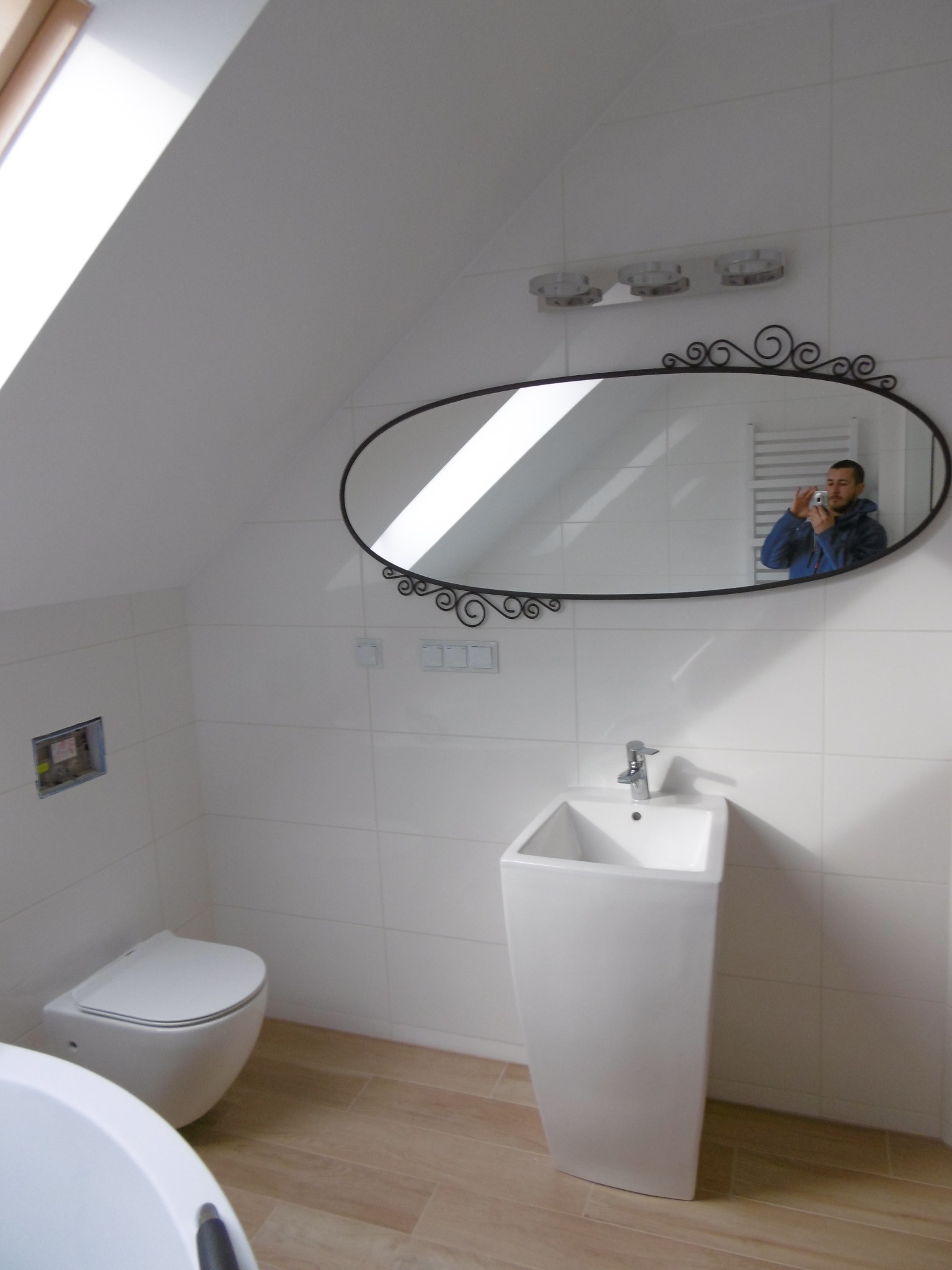 Nowoczesna łazienka na poddaszu z białą ceramiką, podłużnym lustrem z ozdobną ramą, jasnymi płytkami i drewnopodobną podłogą, z widocznym odbiciem fotografa w lustrze.