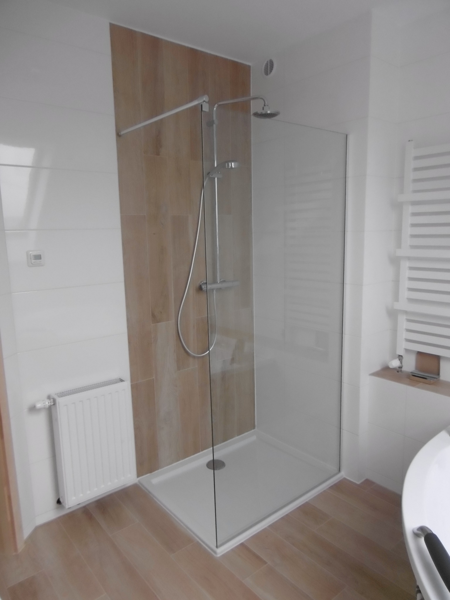 Nowoczesna kabina prysznicowa walk-in z drewnopodobną ścianą i białymi płytkami, widoczny grzejnik drabinkowy i fragment wanny.