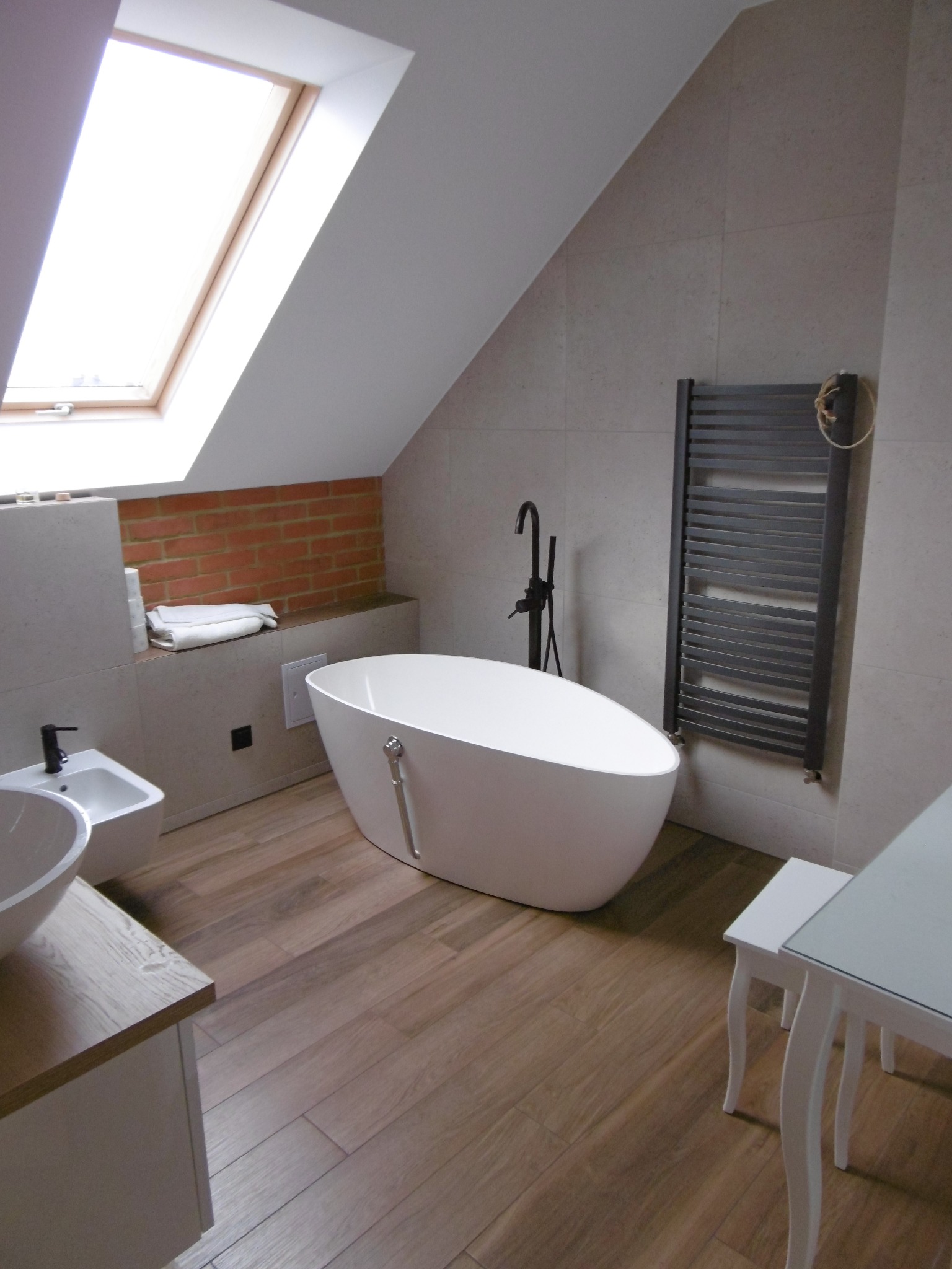 Nowoczesna łazienka na poddaszu z wolnostojącą białą wanną, czarną armaturą, drewnopodobną podłogą, grzejnikiem drabinkowym i oknem dachowym.