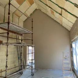 Wnętrze w trakcie prac wykończeniowych: ściany pokryte świeżym tynkiem, widoczne rusztowanie z drabinką, sufit z izolacją i fragmentami pianki montażowej, podłoga z resztkami zaprawy.