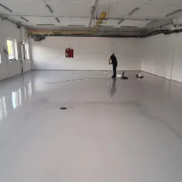 Aplikacja jasnoszarej posadzki w przestronnym, jasnym pomieszczeniu przemysłowym, pracownik wygładza powierzchnię za pomocą specjalistycznego sprzętu.