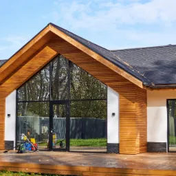 Nowoczesny dom z drewnianą elewacją i dużymi przeszkleniami, widoczny taras z desek i narzędzia budowlane sugerujące prace wykończeniowe.