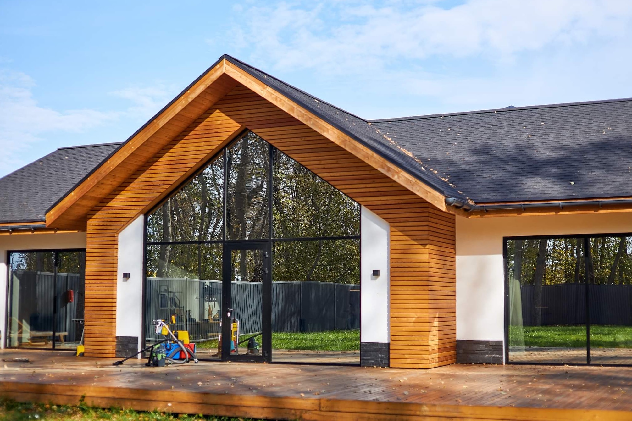 Nowoczesny dom z drewnianą elewacją i dużymi przeszkleniami, widoczny taras z desek i narzędzia budowlane sugerujące prace wykończeniowe.