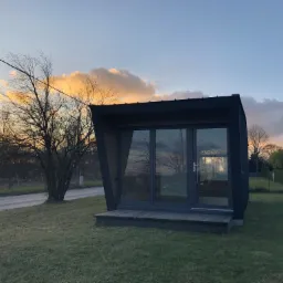 Nowoczesny, czarny domek letniskowy z dużymi przeszkleniami, usytuowany na trawiastym terenie z widokiem na zachodzące słońce i drzewa bez liści.