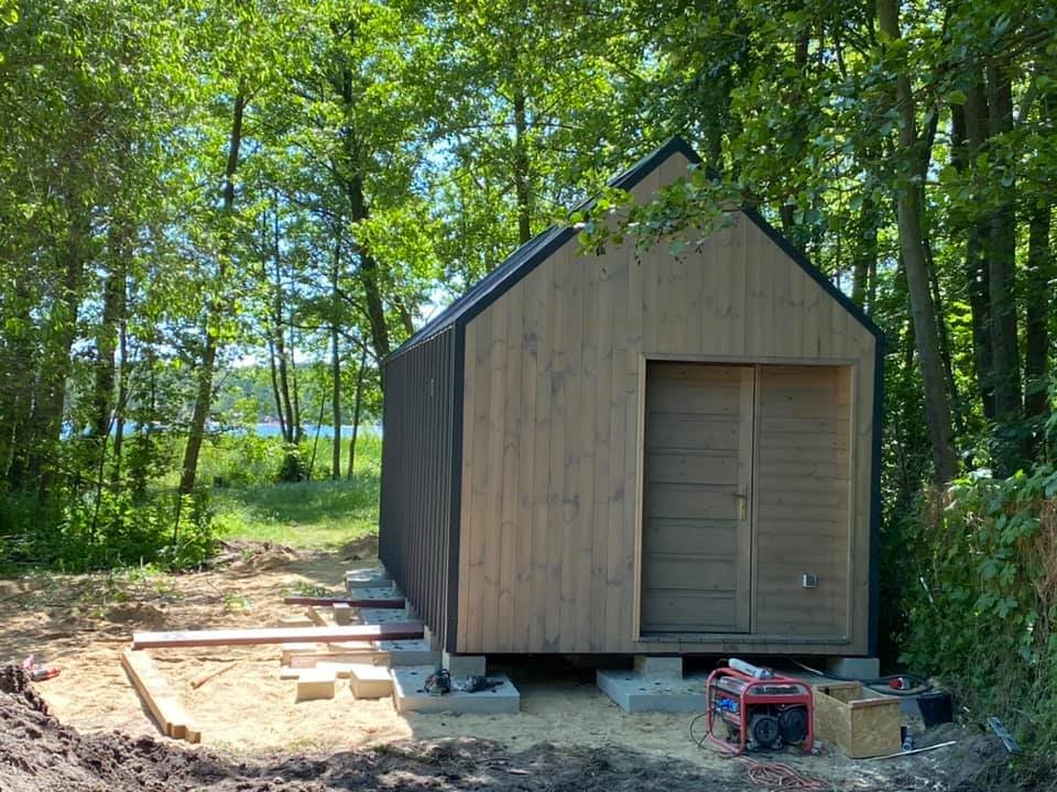 Nieduży, drewniany budynek z dwuspadowym dachem, posadowiony na betonowych bloczkach, widoczny generator prądu i drewniane elementy konstrukcyjne, w tle drzewa i jezioro.