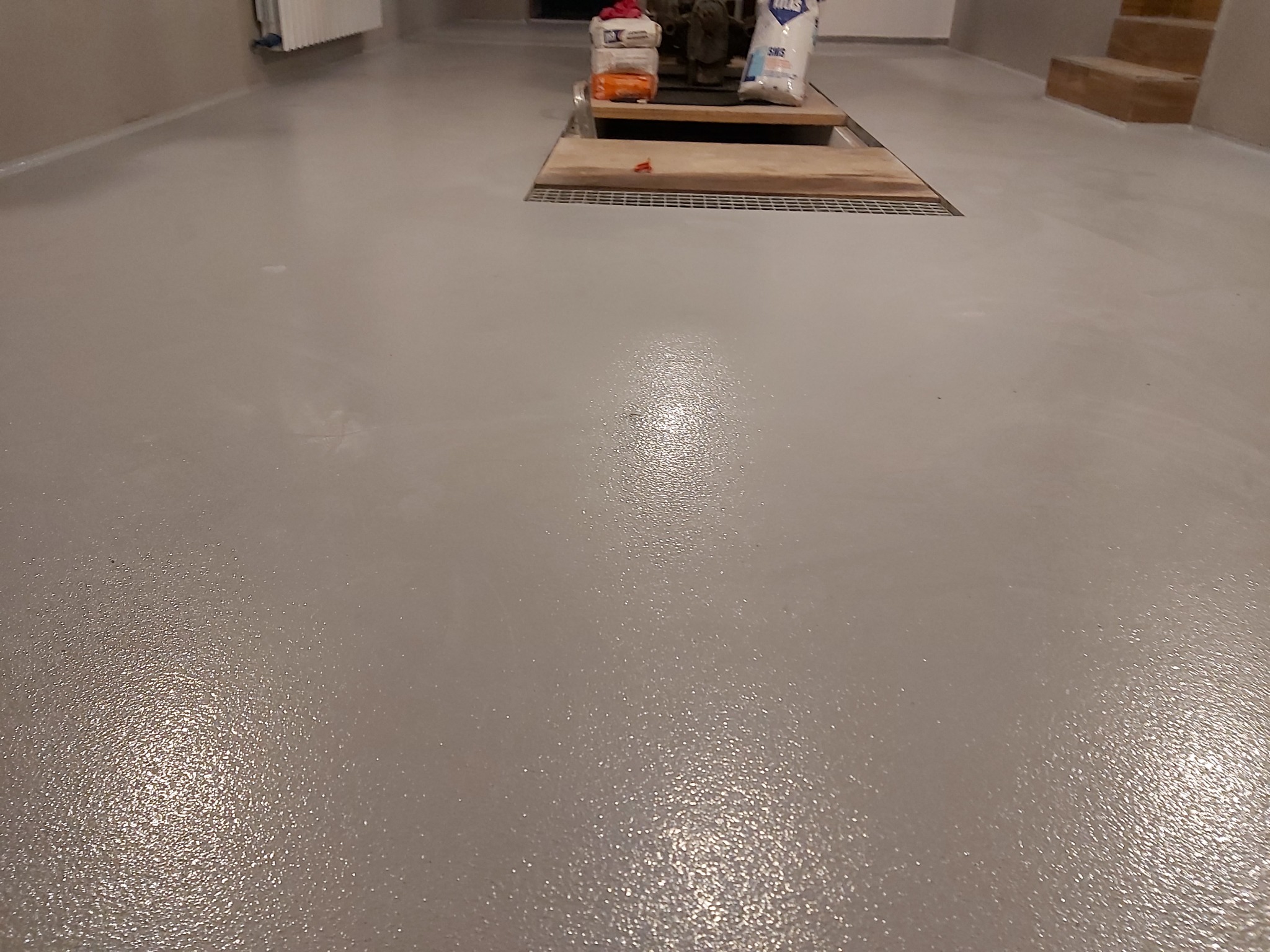 Świeżo wylana, gładka, szara powierzchnia posadzki z widocznym odpływem liniowym i workami z materiałami budowlanymi w tle.