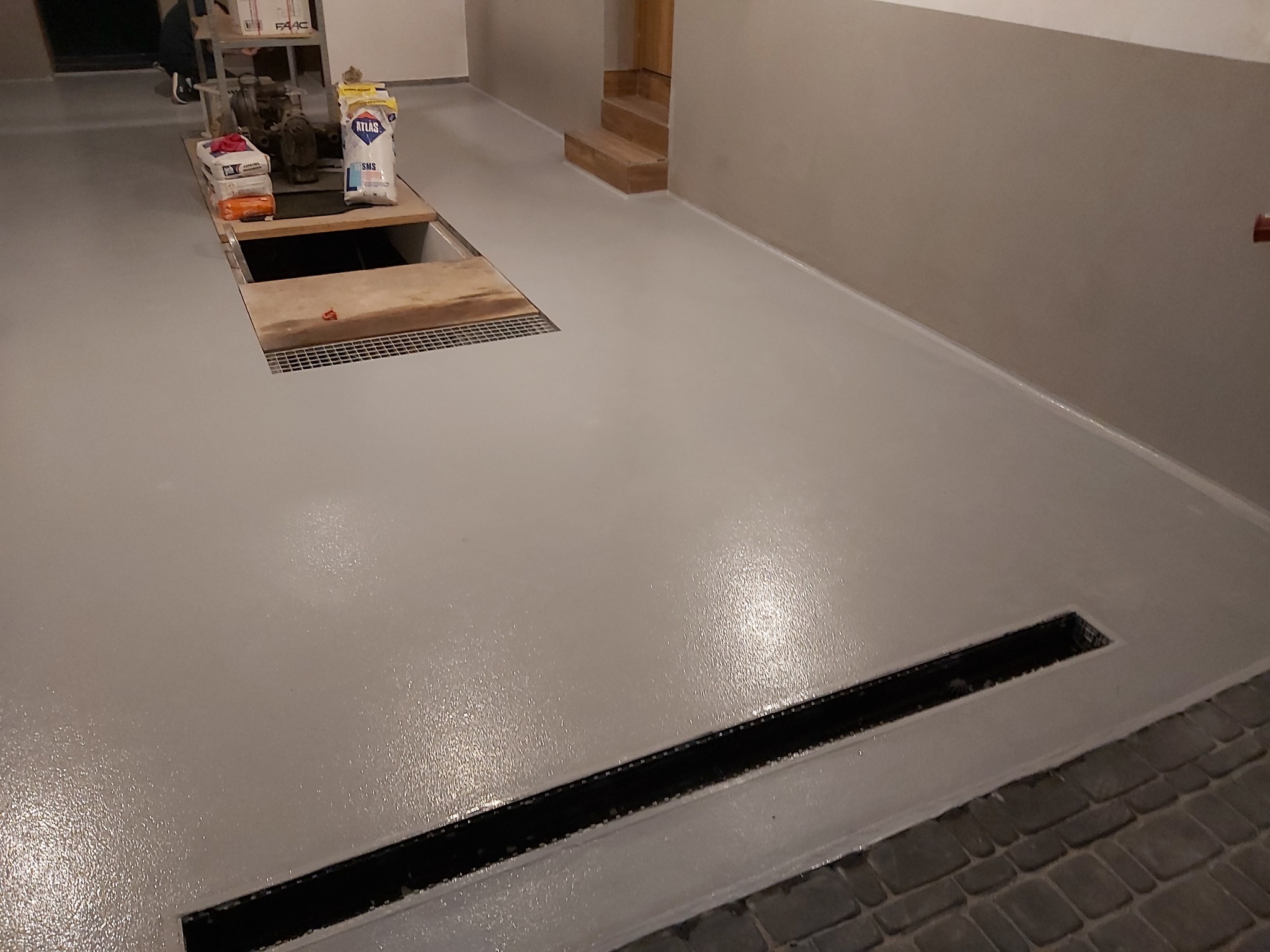 Szara, gładka powierzchnia podłogi z widocznym odpływem liniowym oraz otwartym włazem rewizyjnym z kratką, obok worki z materiałami budowlanymi.