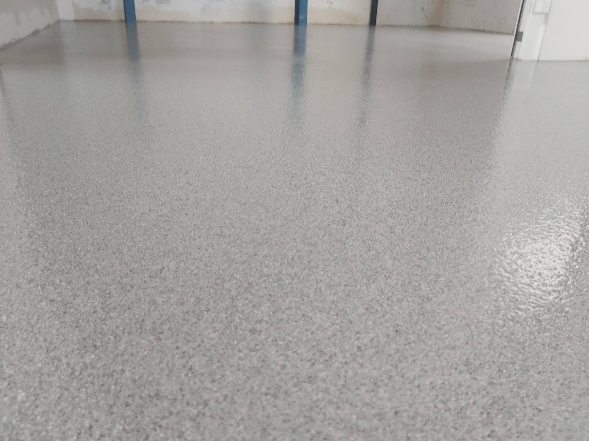 Jasnoszara, gładka posadzka żywiczna z delikatnym ziarnem, świeżo wylana, z widocznymi refleksami światła i fragmentem surowej ściany w tle.
