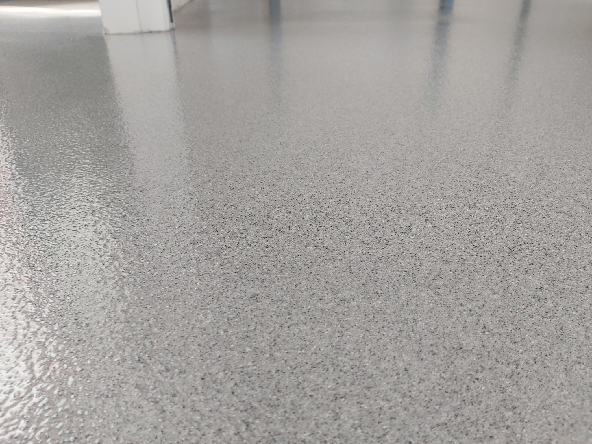 Gładka, szara posadzka z delikatną strukturą, odbijająca światło, w tle fragment białej ściany.