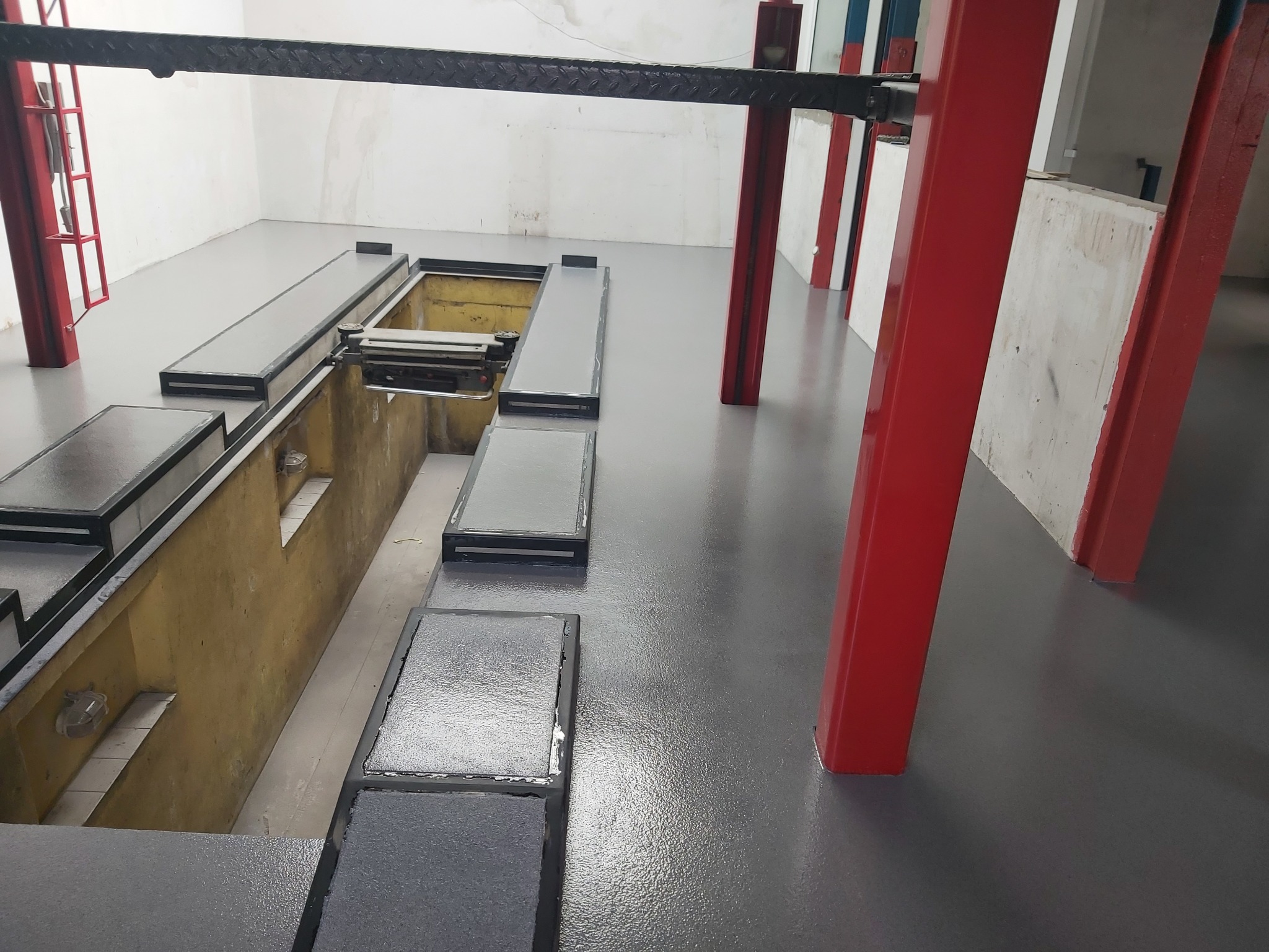 Szara, błyszcząca posadzka w warsztacie samochodowym z kanałem naprawczym i czerwonymi kolumnami podnośnika.