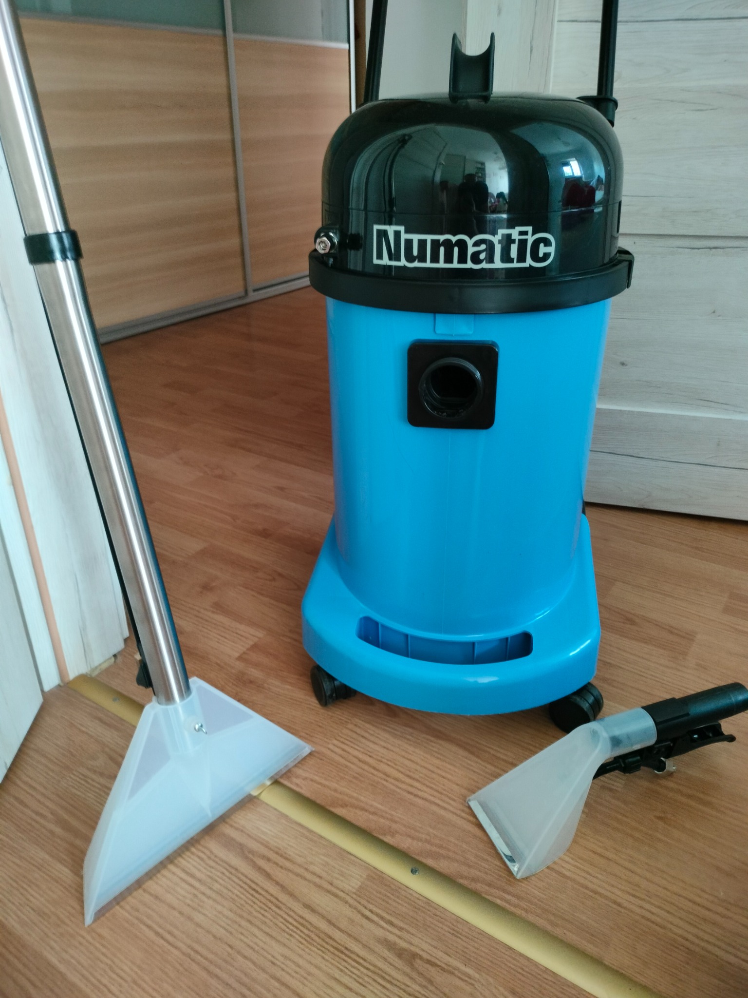 Niebieski odkurzacz piorący marki Numatic z akcesoriami na podłodze z paneli, widoczny wąż i dwie końcówki do czyszczenia tapicerki.