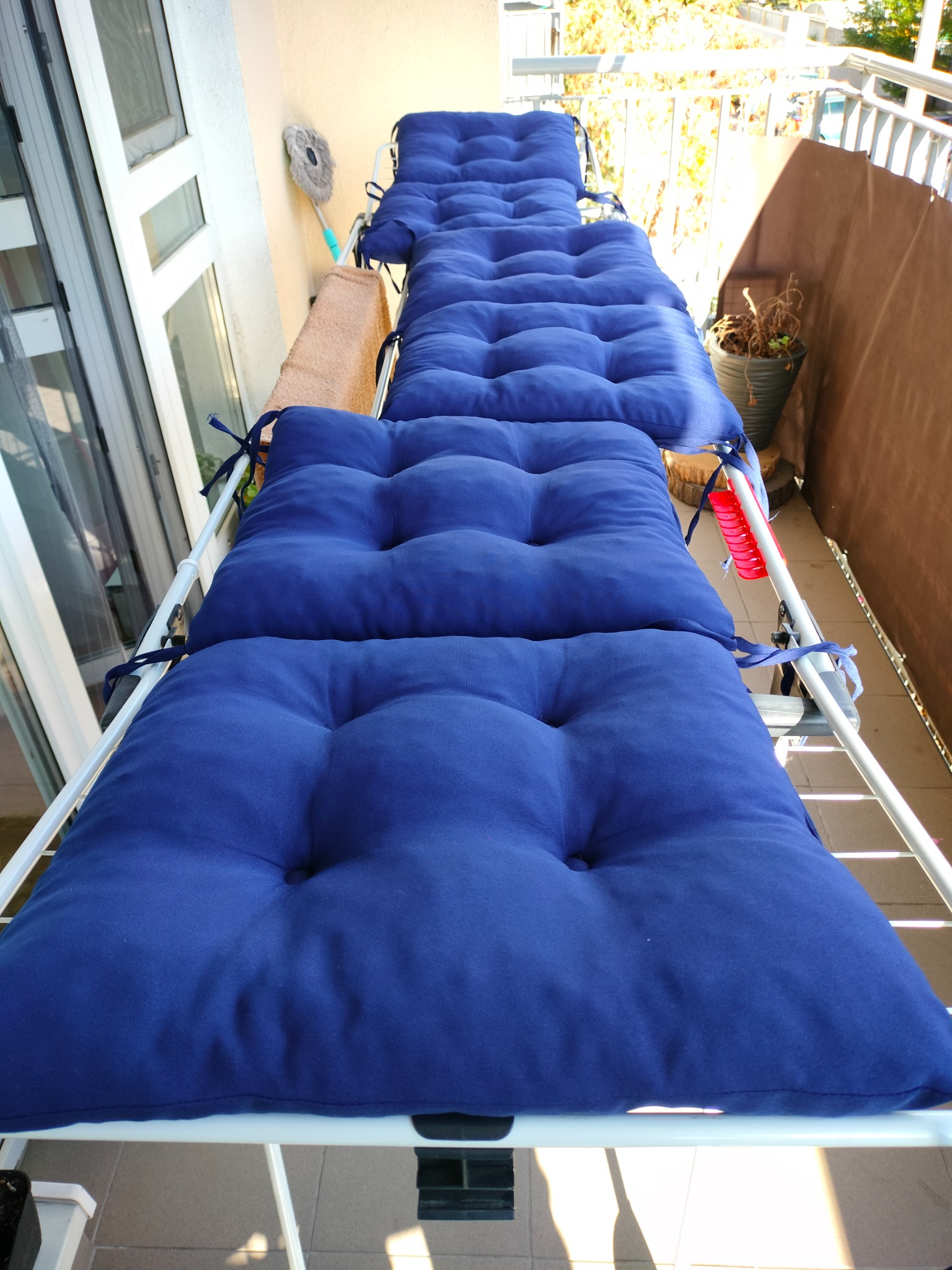 Rząd niebieskich poduszek na metalowej suszarce balkonowej, widok z góry, w tle balkon z widokiem na drzewa.