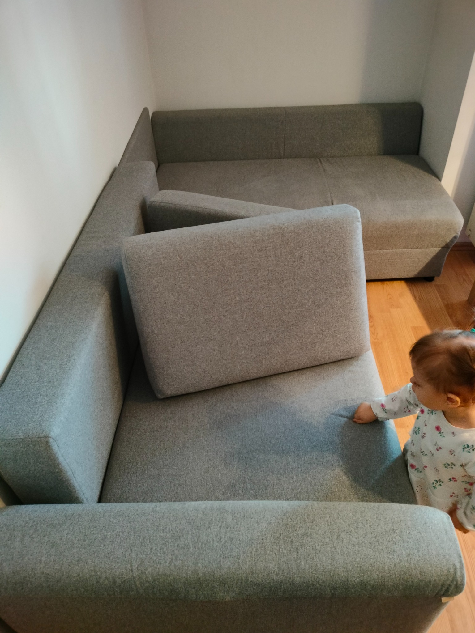 Szara, narożna kanapa tapicerowana z poduszką, widziana z góry, z małym dzieckiem w kwiecistej piżamie stojącym obok.