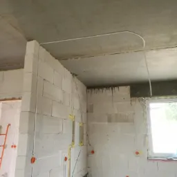 Surowy stan budynku w trakcie prac instalacyjnych: widoczne kable elektryczne prowadzone po ścianach z betonu komórkowego, puszki instalacyjne w kolorze pomarańczowym, oraz kabel pod sufitem.