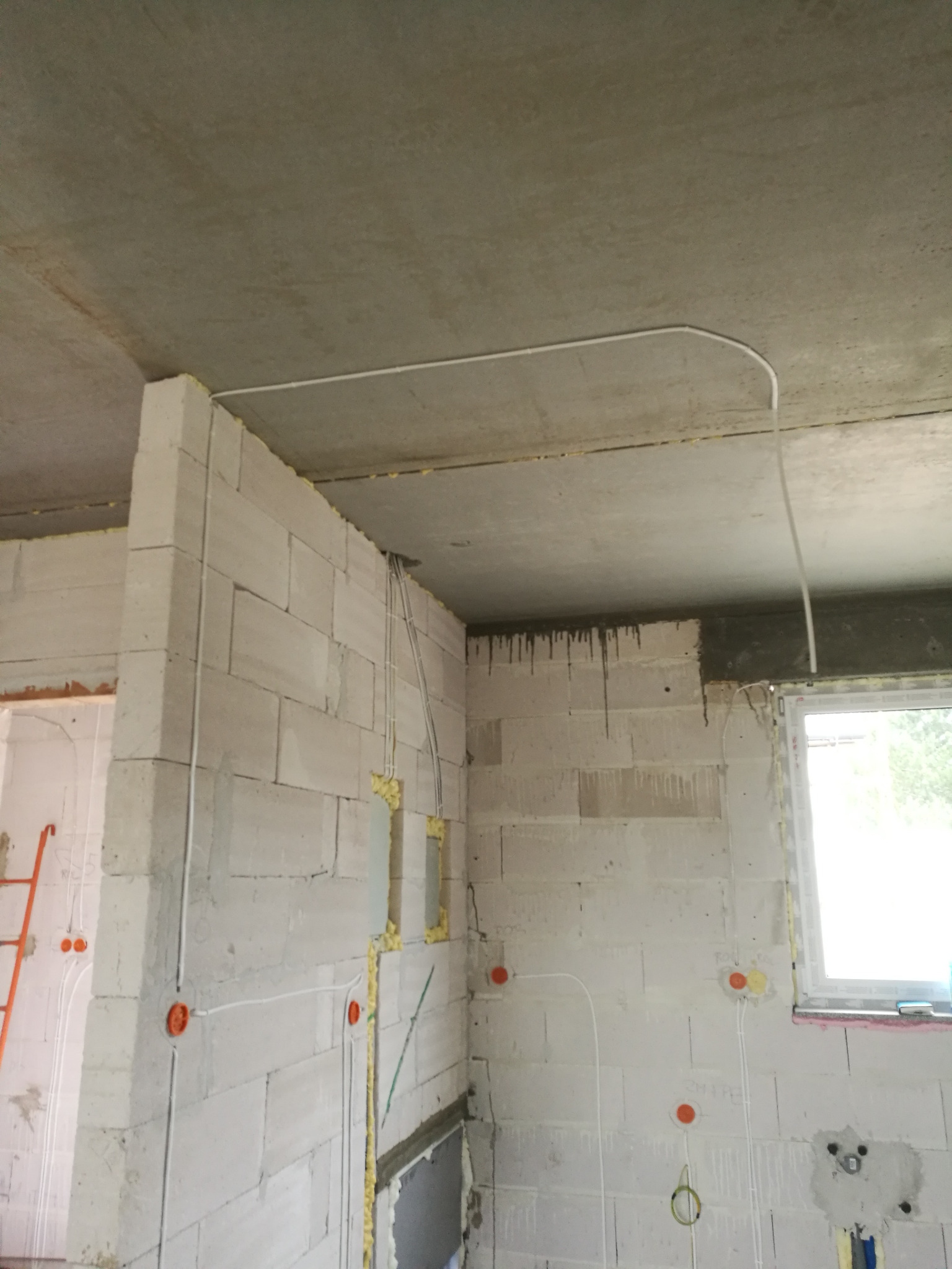 Surowy stan budynku w trakcie prac instalacyjnych: widoczne kable elektryczne prowadzone po ścianach z betonu komórkowego, puszki instalacyjne w kolorze pomarańczowym, oraz kabel pod sufitem.