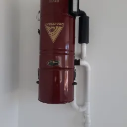 Ciemnoczerwony agregat centralnego odkurzacza Cyclo Vac H725 z filtrem i białymi rurami PCV zamontowany na białej ścianie.