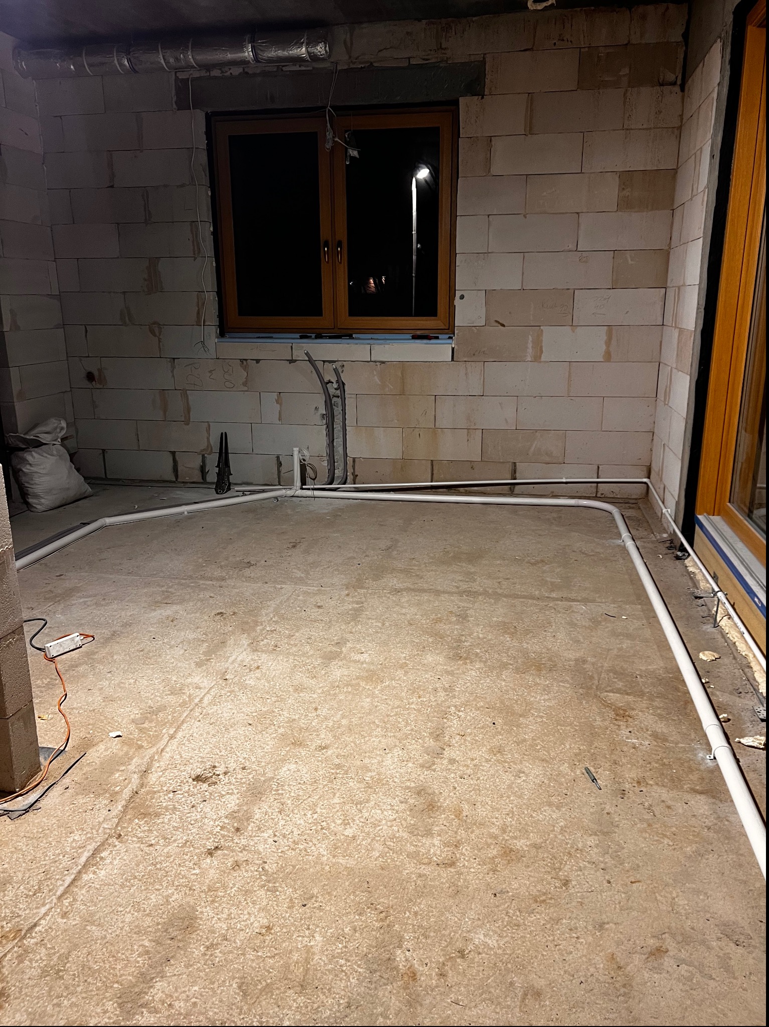 Surowy stan podłogi w pomieszczeniu z instalacją rur PCV przy ścianach z bloczków, widoczne okno i worek z materiałem budowlanym.