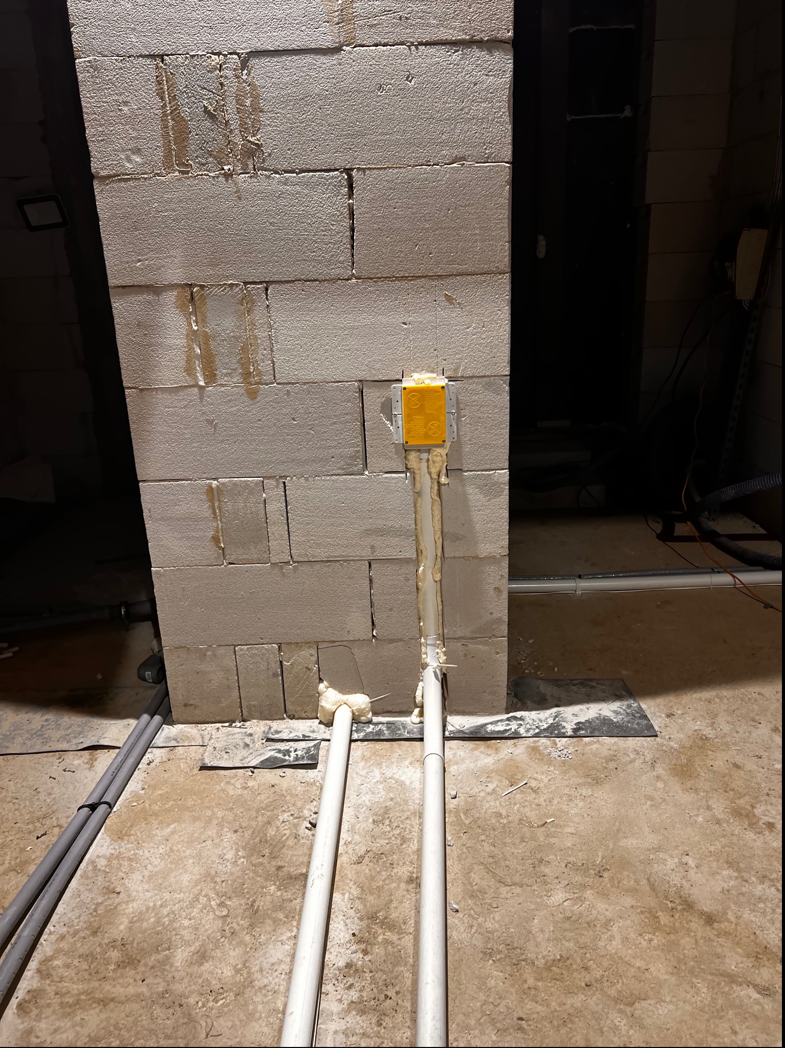 Surowy mur z betonu komórkowego z żółtą puszką instalacyjną i rurami do odkurzacza centralnego zabezpieczonymi pianką montażową, widoczne elementy instalacji na surowym podłożu.