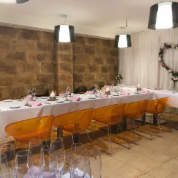 Uroczysta sala z długim stołem nakrytym białym obrusem, różowe serwetki, przezroczyste pomarańczowe krzesła, ściana z cegieł i dekoracja w kształcie serca z kwiatów.