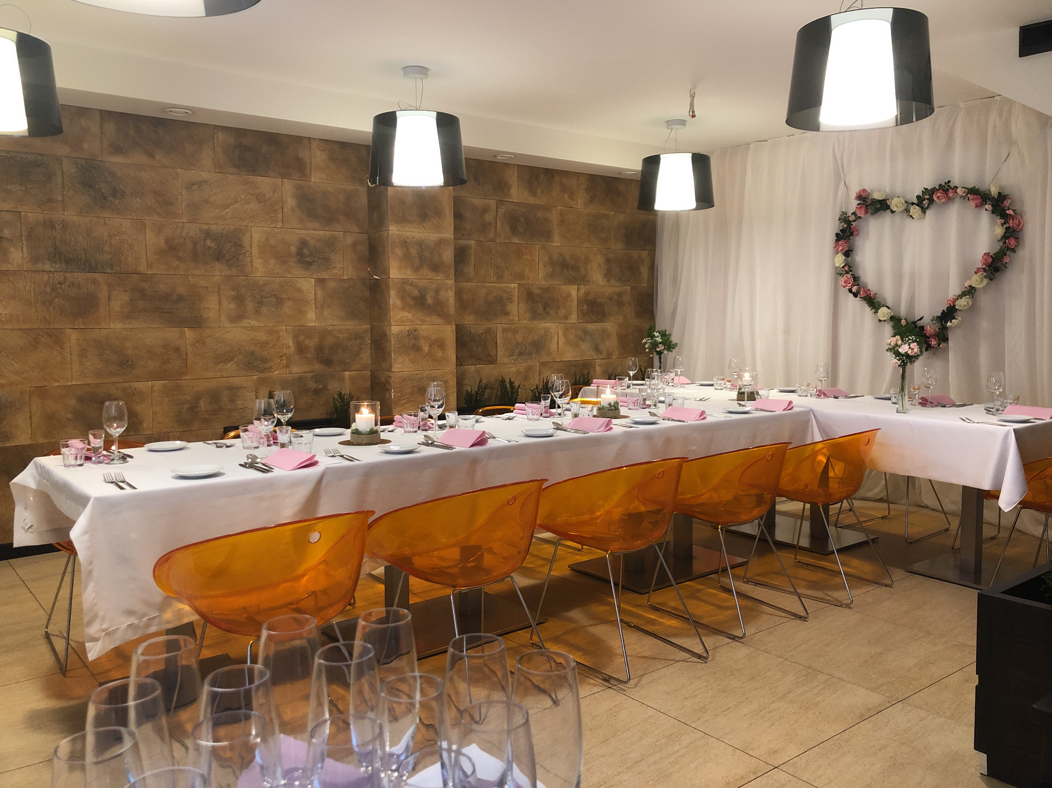 Uroczysta sala z długim stołem nakrytym białym obrusem, różowe serwetki, przezroczyste pomarańczowe krzesła, ściana z cegieł i dekoracja w kształcie serca z kwiatów.