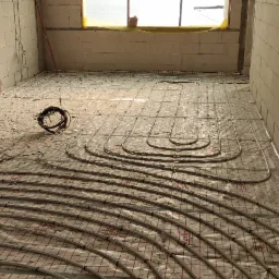 Rozłożone rury ogrzewania podłogowego na folii z siatką w surowym pomieszczeniu z białych bloczków, zwinięty przewód na podłodze.