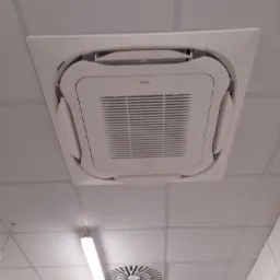 Sufitowa jednostka wewnętrzna marki Daikin z widoczną kratką wentylacyjną i oświetleniem liniowym na białym suficie kasetonowym.