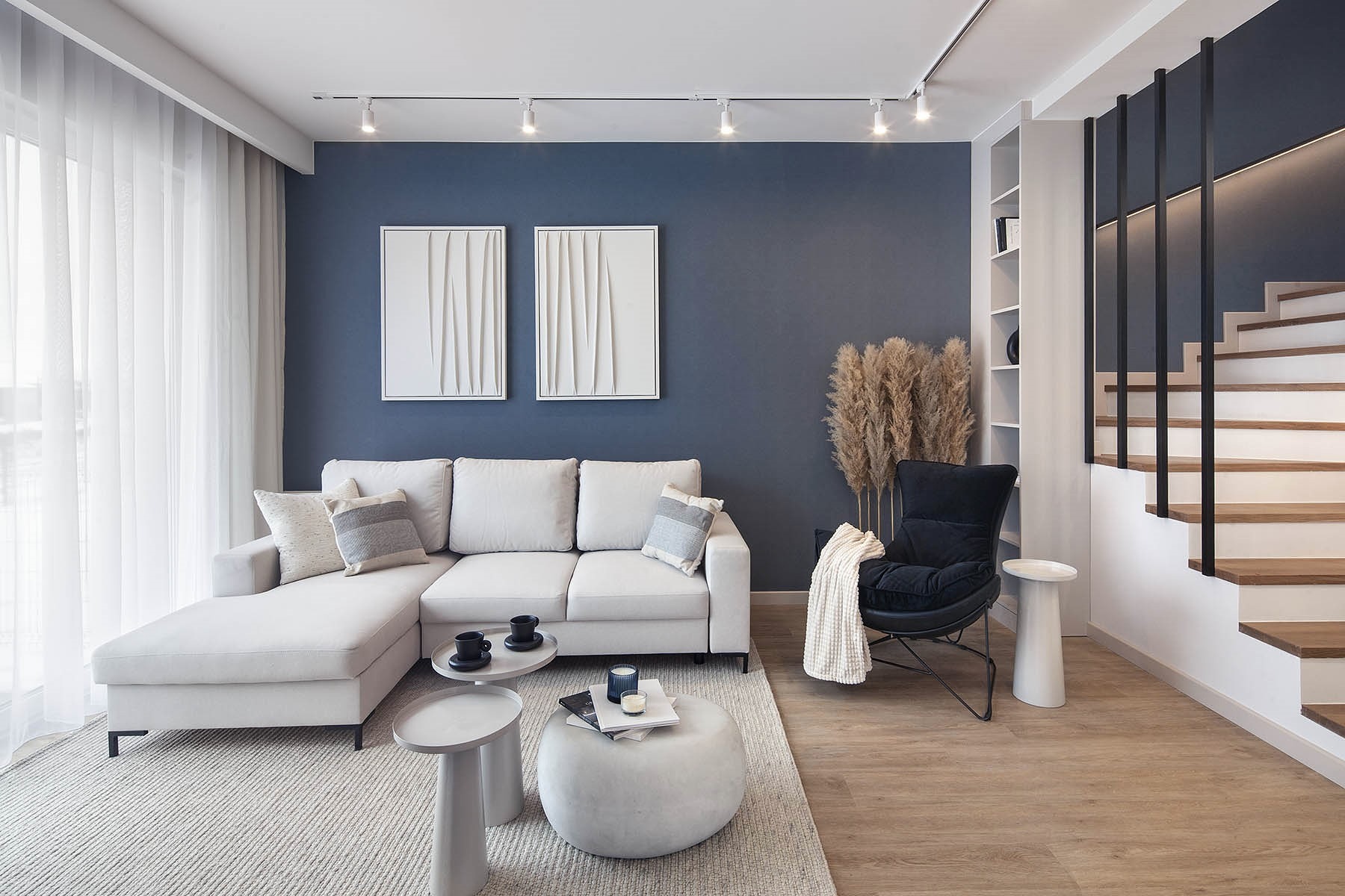 Stylowe wnętrze: narożna sofa w odcieniach szarości, dekoracyjne obrazy na granatowej ścianie, drewniane schody z czarną balustradą i pampasowa trawa.