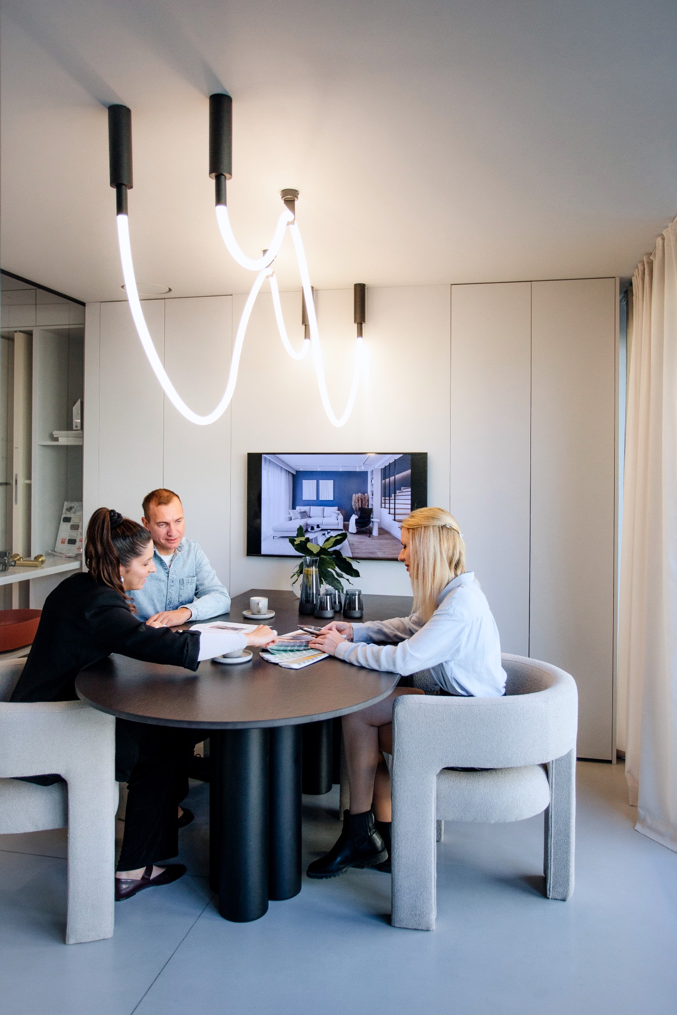 Spotkanie projektowe: dwie kobiety i mężczyzna omawiają paletę kolorów przy okrągłym stole w nowoczesnym wnętrzu z designerską lampą i telewizorem prezentującym wizualizację.