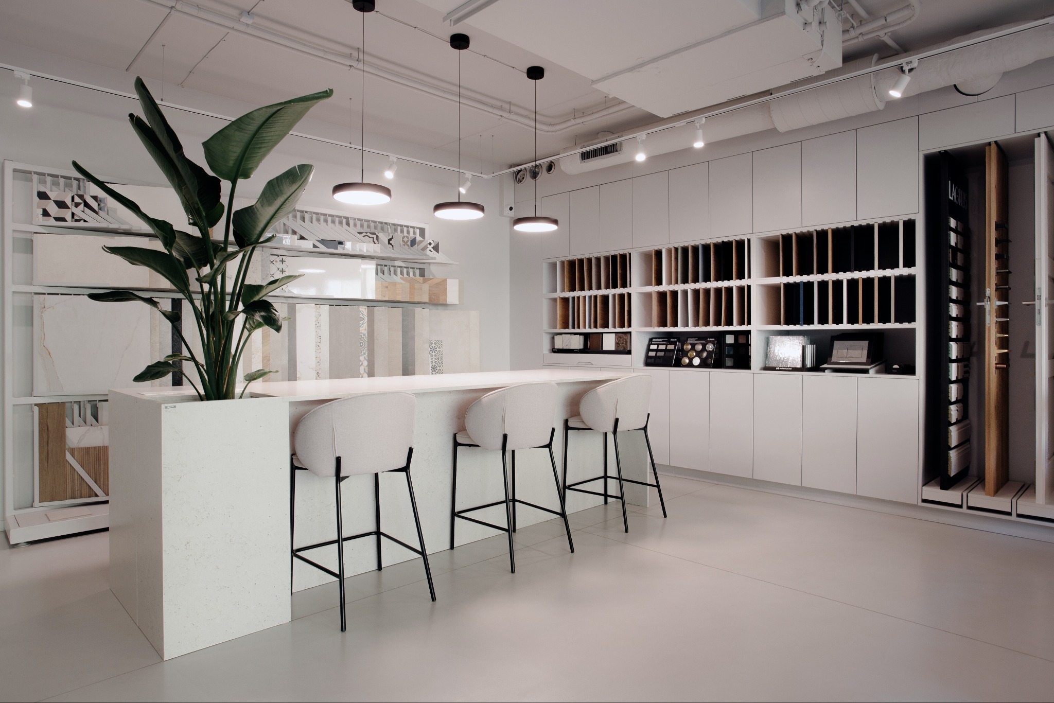 Jasne wnętrze sklepu z płytkami: minimalistyczny design, wysokie krzesła barowe, ekspozycja próbek płytek, oświetlenie punktowe i donica z zieloną rośliną.