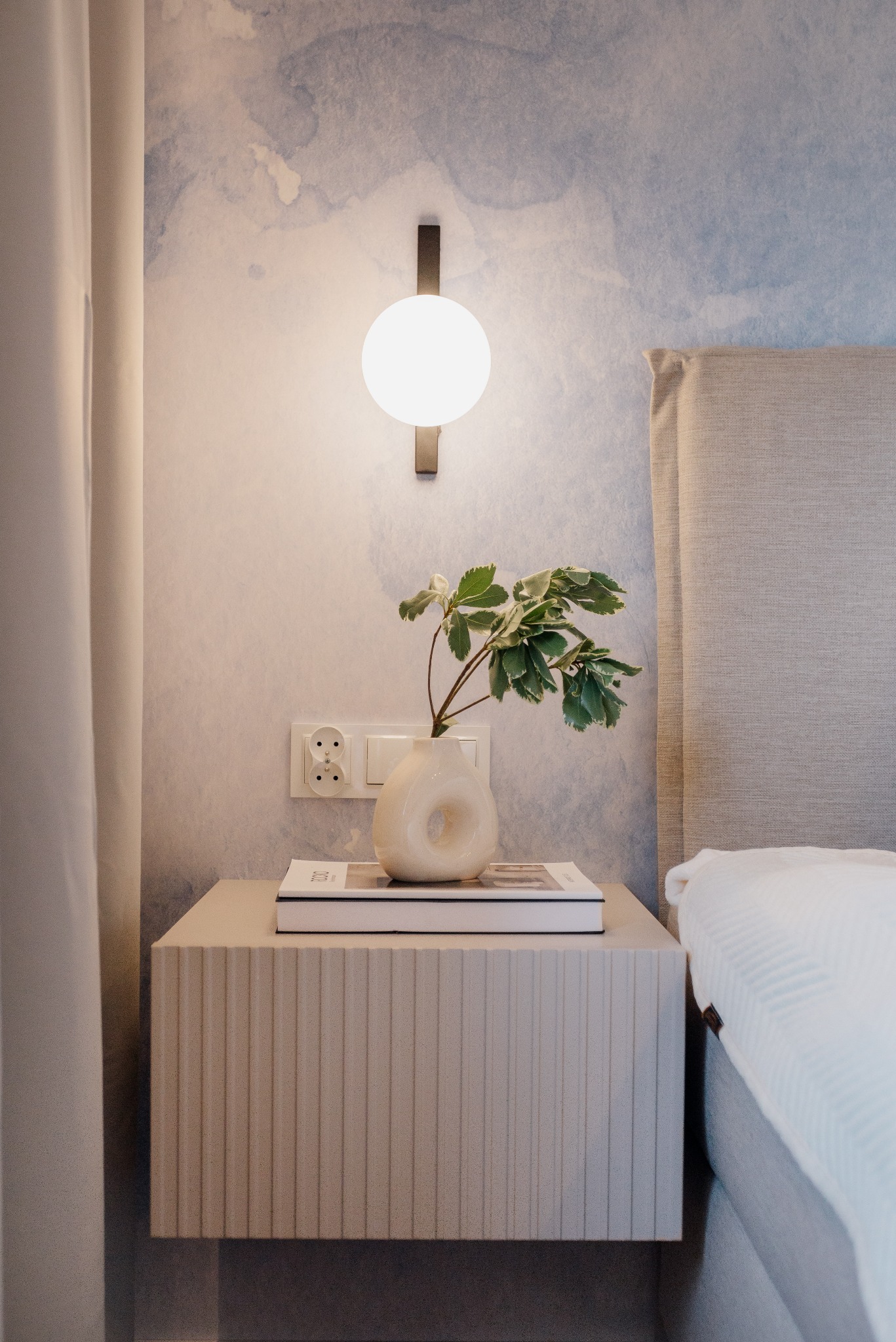 Sypialnia: lampa ścienna nad szafką nocną z wazonem i książkami, tapeta w odcieniach błękitu, fragment łóżka z szarym zagłówkiem. Minimalistyczny design.