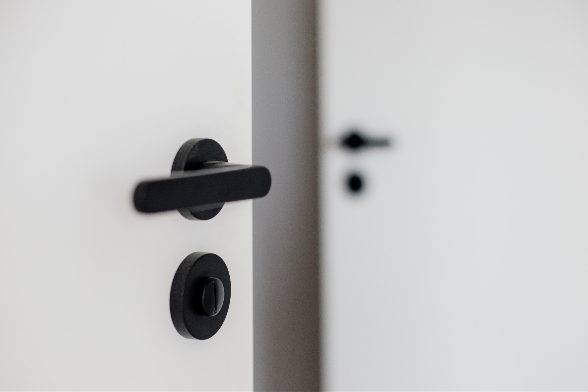 Białe drzwi z czarną, minimalistyczną klamką i zamkiem. W tle widoczne drugie drzwi z podobną klamką, tworzące spójny, nowoczesny design wnętrza.