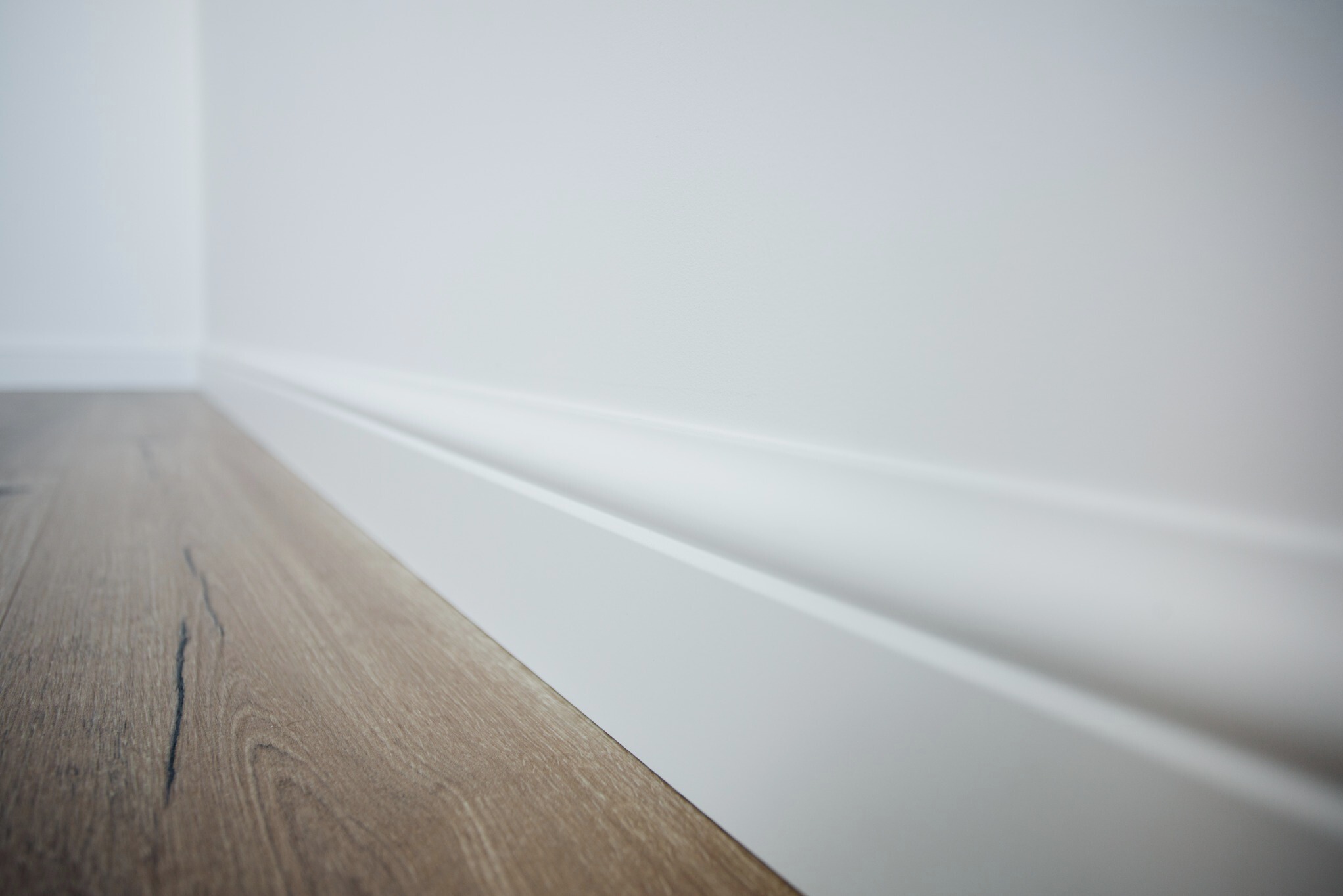 Jasne panele podłogowe z drewnianym wzorem, biała listwa przypodłogowa przy białej ścianie, widok z niskiej perspektywy, oświetlenie naturalne.