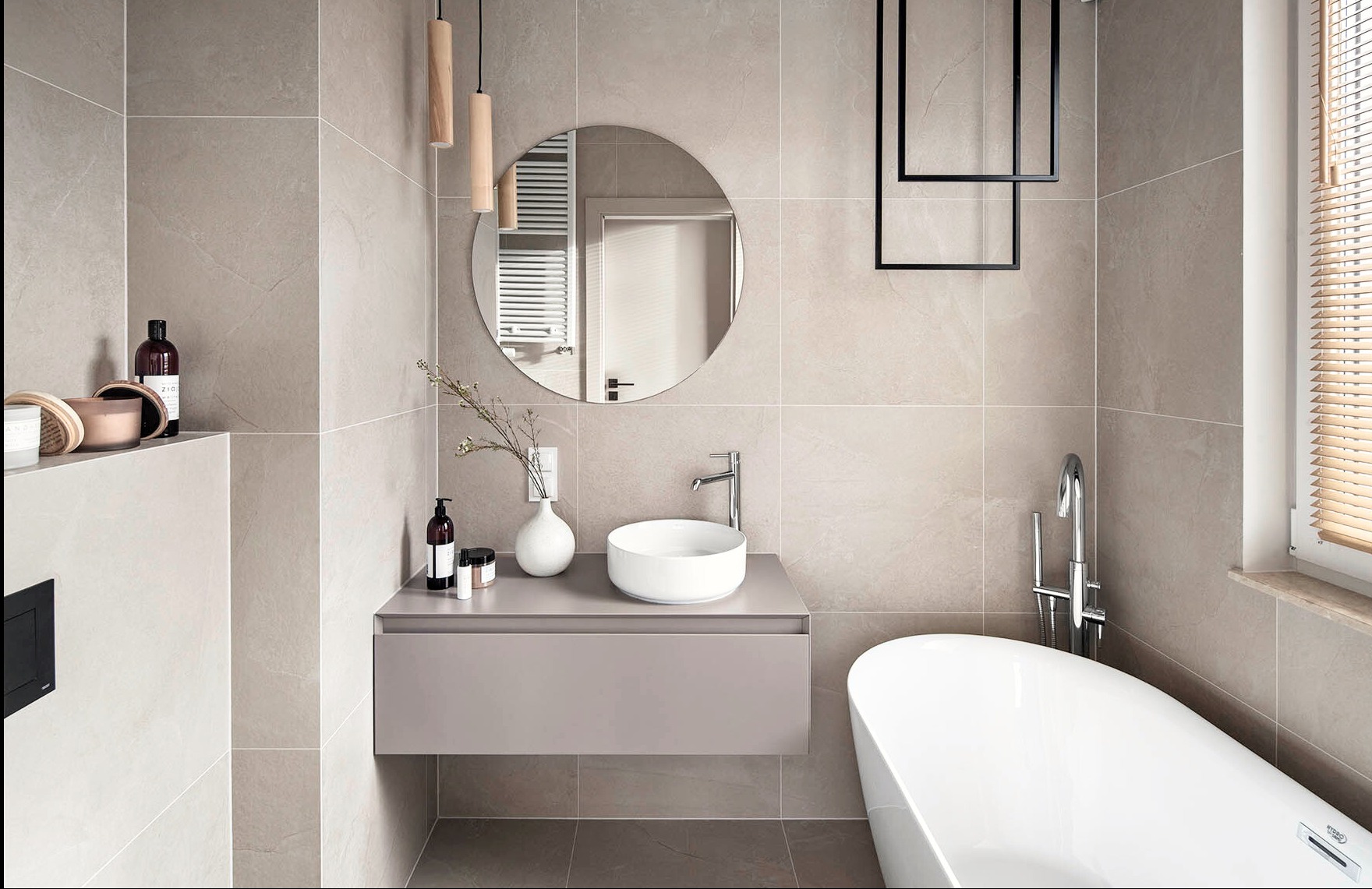 Beżowa łazienka z okrągłym lustrem, wiszącą szafką z umywalką i wolnostojącą wanną. Minimalistyczne oświetlenie i płytki imitujące kamień.