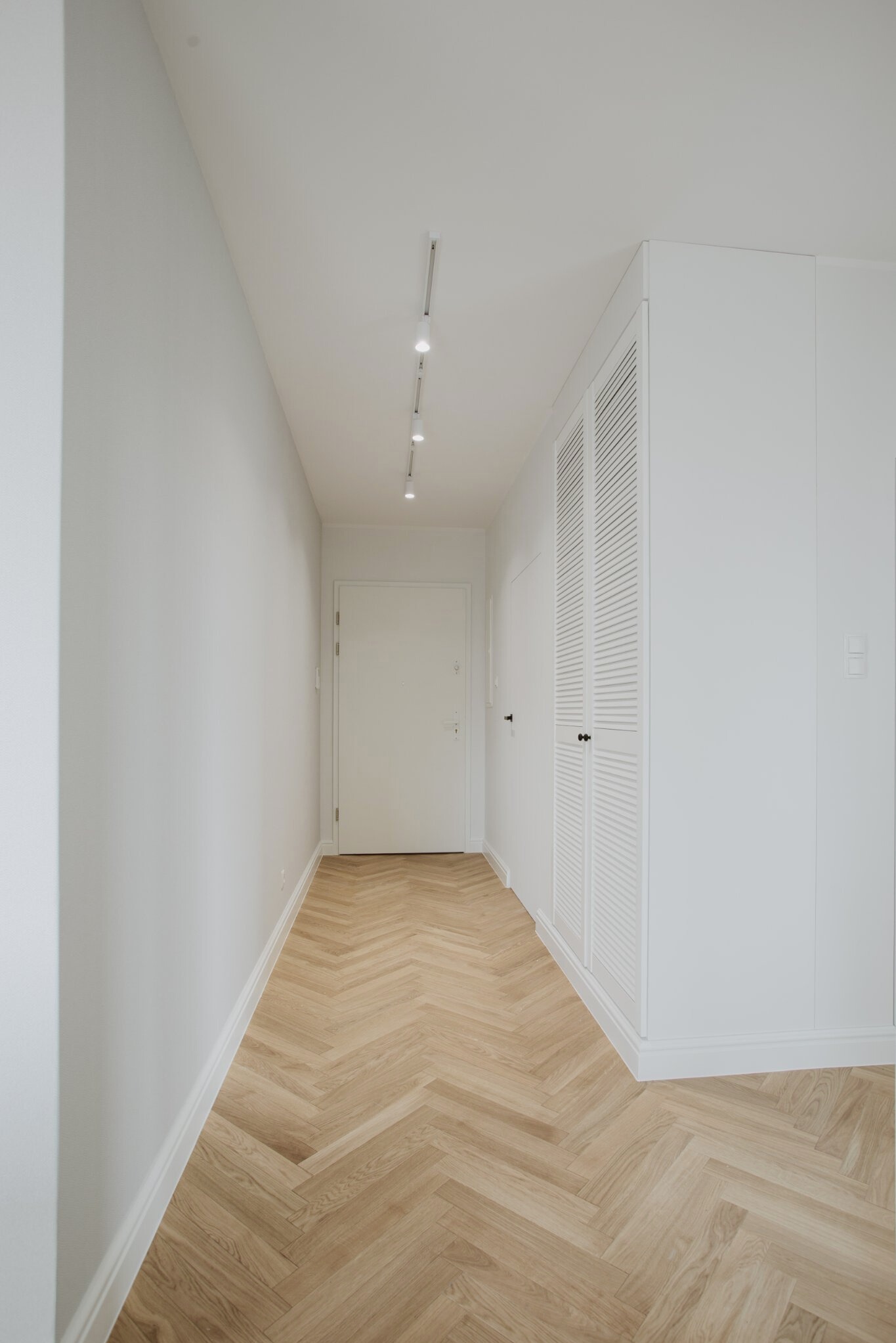 Jasny korytarz z parkietem ułożonym we wzór jodełki, oświetleniem punktowym i zabudowaną szafą z lamelowymi drzwiami. Minimalistyczny design.
