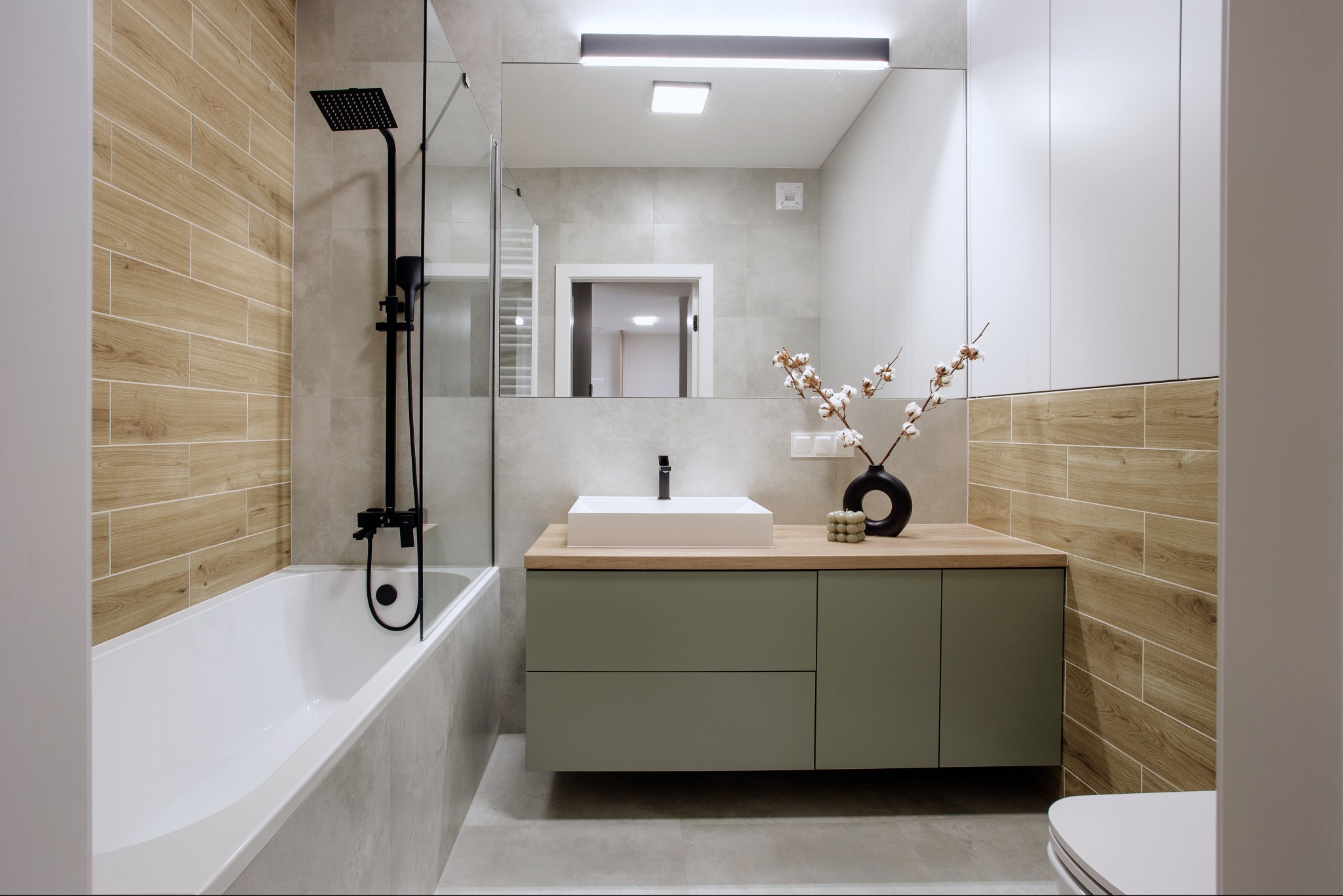 Nowoczesna łazienka z wanną, czarną armaturą i umywalką na szafce w kolorze oliwkowym. Drewnopodobne płytki na ścianie i minimalistyczny design.