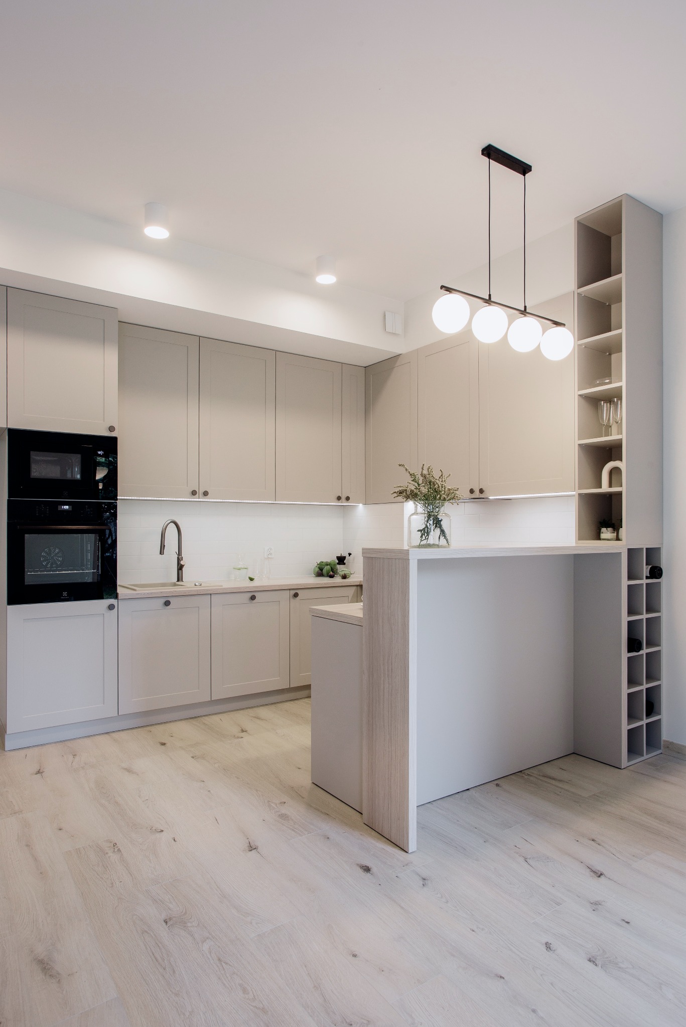 Jasna kuchnia w stylu skandynawskim z wyspą, oświetleniem LED pod szafkami i drewnianą podłogą. Nowoczesny design z elementami dekoracyjnymi, jak wazon z kwiatami i półka na wino.