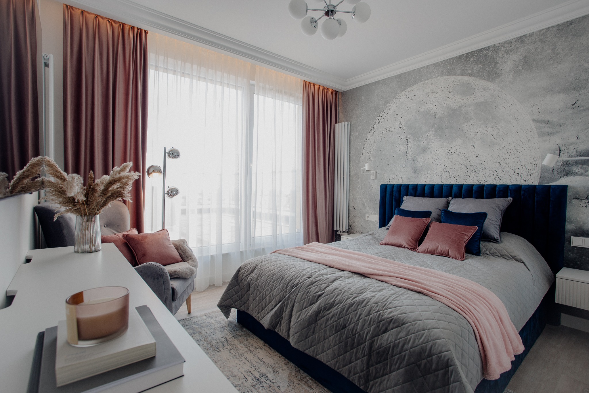 Stylowa sypialnia z łóżkiem w odcieniach szarości i granatu, fototapeta z motywem księżyca, zasłony w kolorze bordo i dekoracyjny fotel z pampasami.