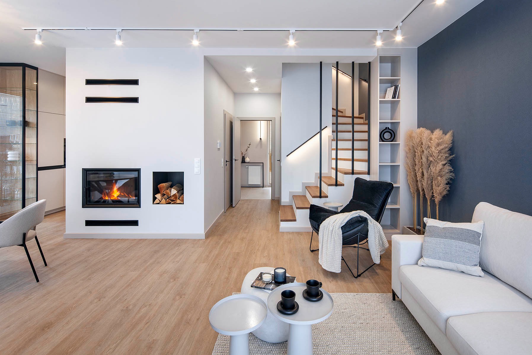 Nowoczesny salon z kominkiem, drewnianymi schodami i oświetleniem punktowym. Minimalistyczny design z drewnianą podłogą i dekoracjami z pampasów.