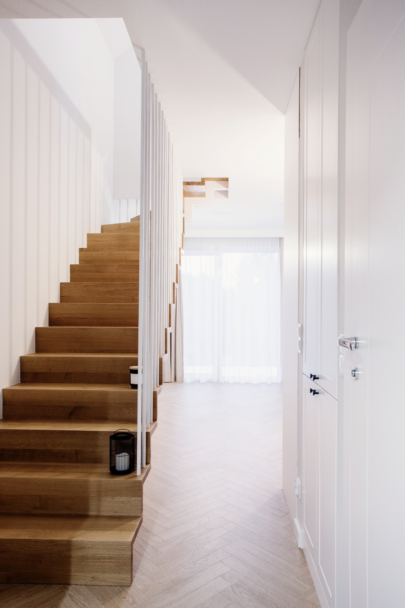 Jasne wnętrze domu: drewniane schody z białymi balustradami, parkiet ułożony w jodełkę, biała szafa wnękowa. Minimalistyczny design.