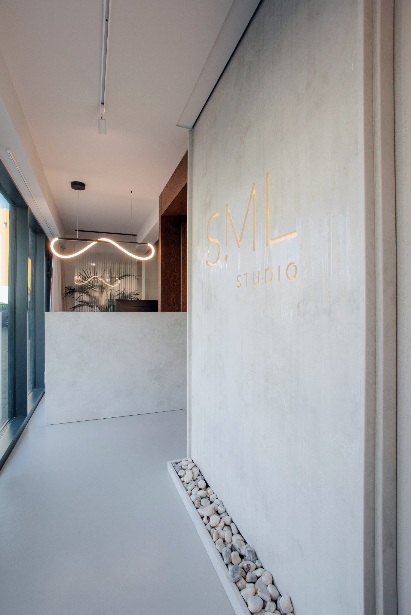 Minimalistyczne wnętrze studia z recepcją, oświetleniem LED i ścianą z subtelnym logo 'S.M.L. STUDIO' oraz dekoracyjnym elementem z białych kamieni przy podłodze.
