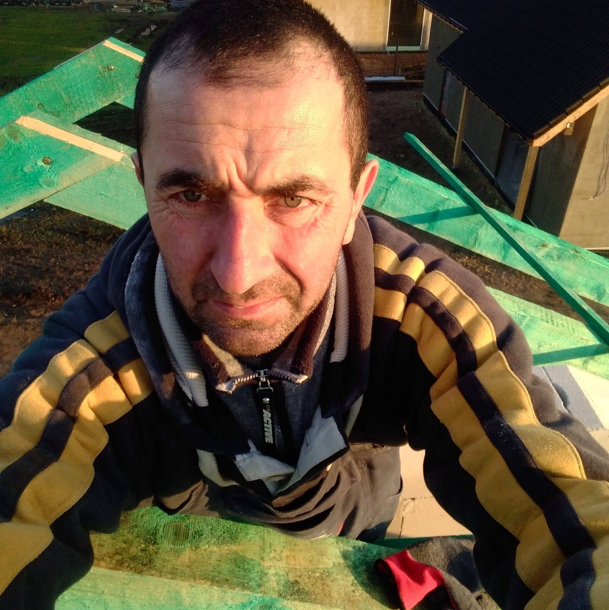 Autoportret mężczyzny na tle konstrukcji dachu z pomalowanych na zielono belek, w tle budynek z czarnym dachem.