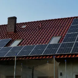 Panele słoneczne na czerwonym dachu domu jednorodzinnego z ceglanym kominem, widoczne okna dachowe i siatka ochronna w dolnej części paneli.