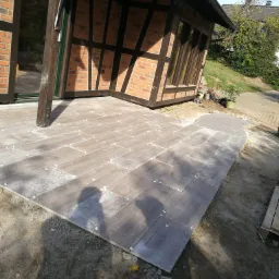 Układanie brązowych płytek imitujących drewno na zewnątrz budynku z cegły, widoczne krzyżyki dystansowe i fragment elewacji.