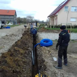 Wykop pod instalacje w trakcie prac ziemnych; widoczny wąski rów, ułożone rury instalacyjne, pracownicy, mini koparka i domy w tle.