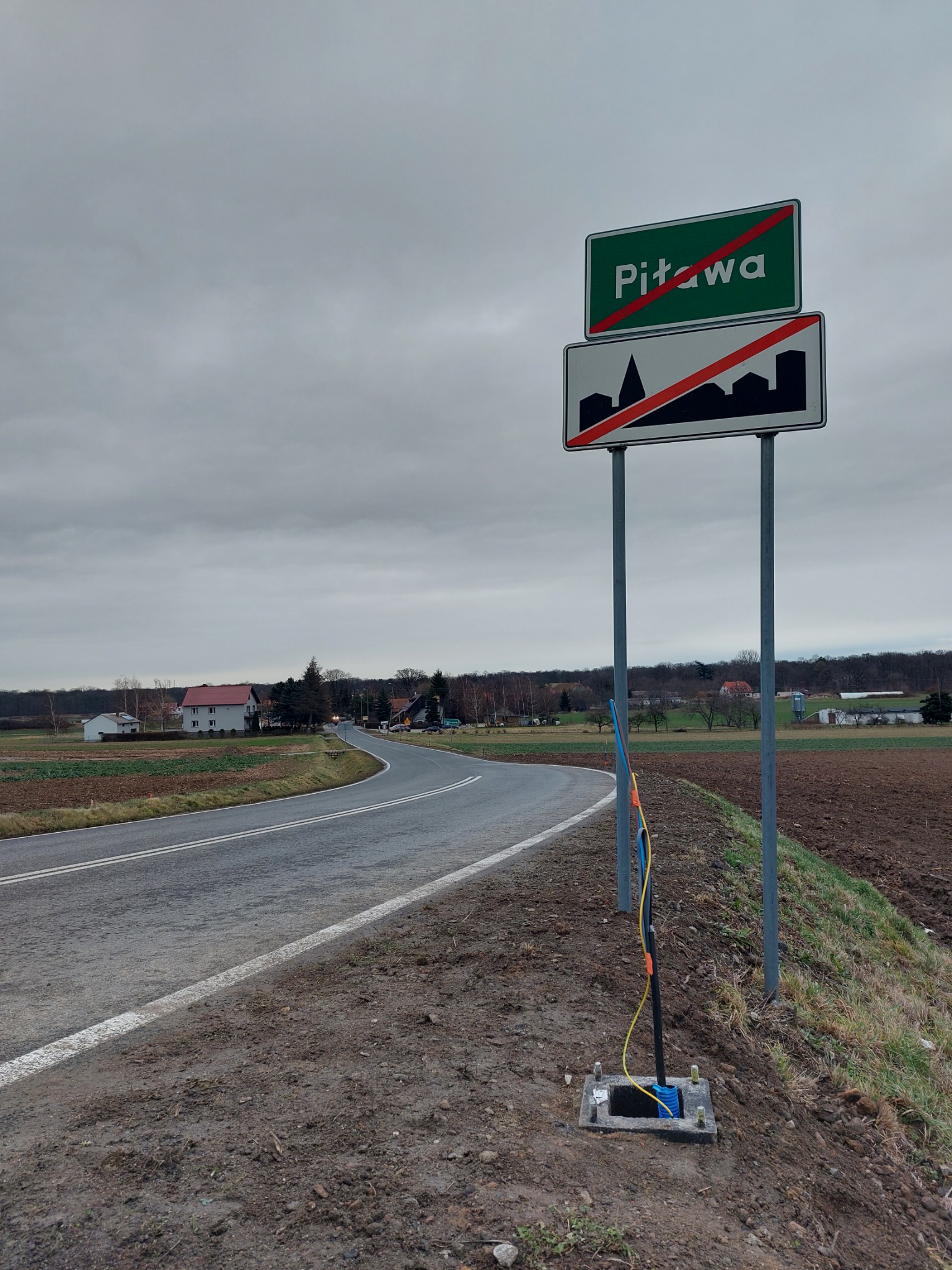 Zakończone prace w Gogolinie: Wprowadzenie kabla światłowodowego do studzienki telekomunikacyjnej przy drodze, widoczny znak drogowy 'Piława' przekreślony czerwoną linią.