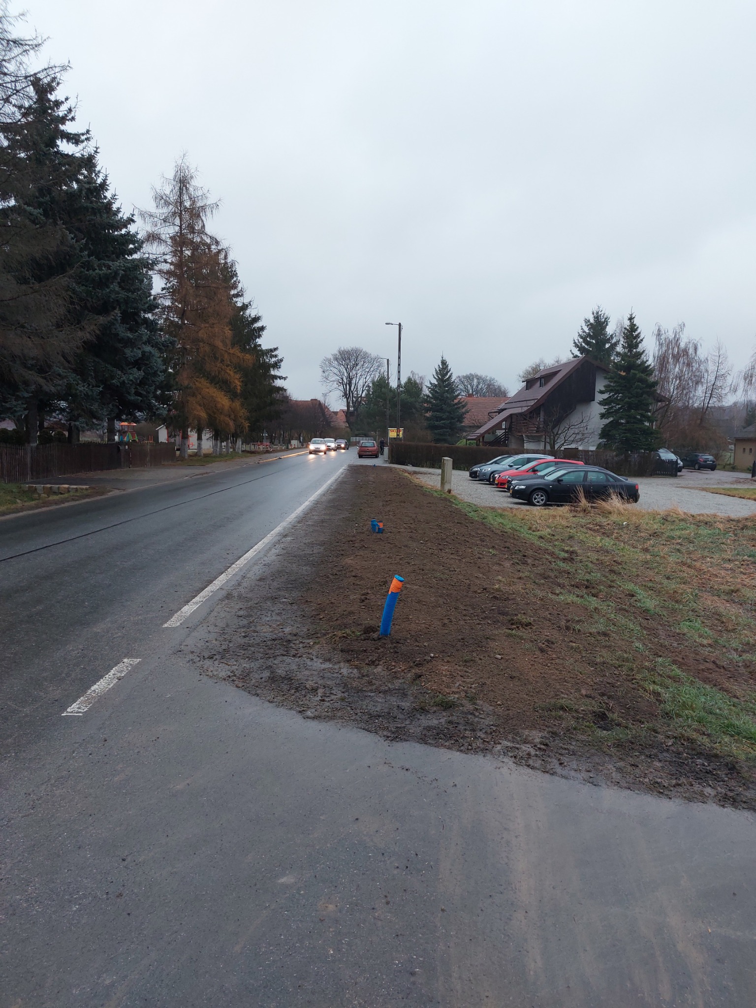 Ulica po deszczu z widocznymi, wystającymi rurami oznaczającymi miejsce przewiertu sterowanego, w tle zaparkowane samochody i domy.