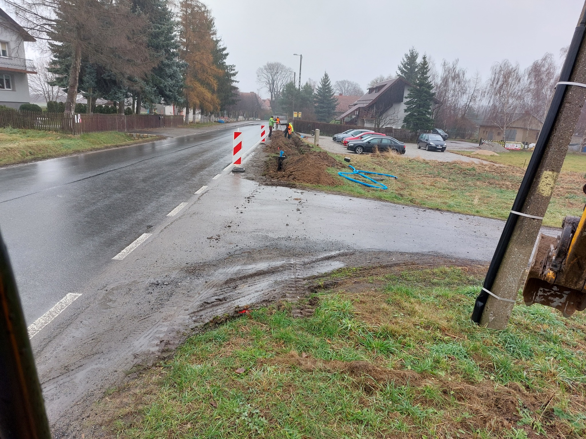 Wykonanie wykopu wzdłuż drogi, widoczni pracownicy w odblaskowych kamizelkach, zabezpieczenia drogowe oraz niebieskie rury na trawie.